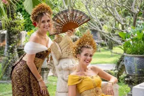 Two women wearing Balinese traditional costumes and gold headdresses posing beside a stone statue