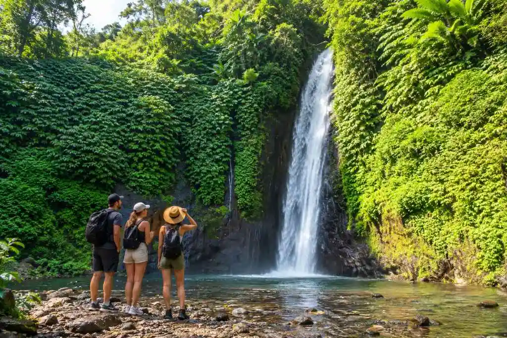Bali Nature Tour featuring three travelers admiring a tall waterfall surrounded by lush tropical greenery in Bali