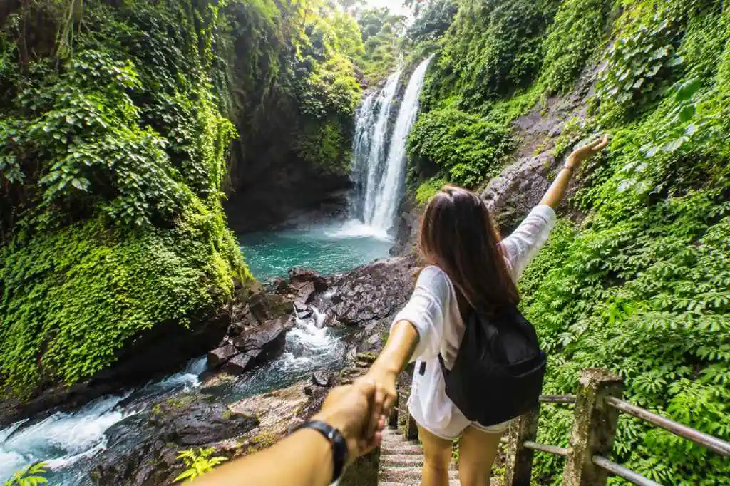 Traveler exploring a tall Sambangan waterfall with one arm raised beside a lush green cliff