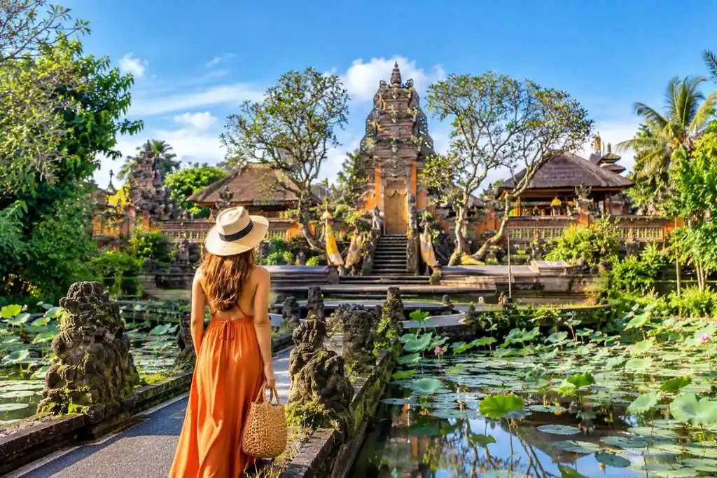 Woman in a white summer dress and straw hat walking toward a traditional Balinese temple surrounded by lotus ponds during an Ubud Tour.